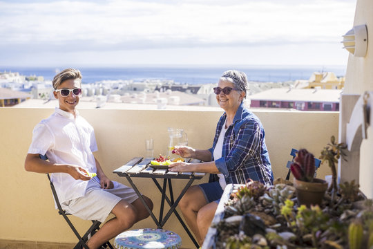 happy leisure activity on the terrace rooftop having breakfast with smiles and happiness for grandmother and teenager family caucasian people. ocean and buildings view, outdoor coule together