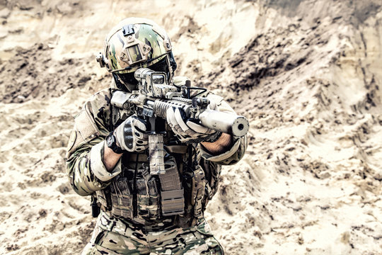 Special operations forces fighter, modern infantry soldier in full ammunition, aiming with red dot sight and shooting with assault carbine in desert area. Fire contact with enemy during war conflict