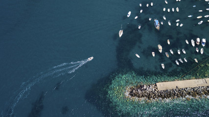 Perpendicular aerial view of a white motorboat entering a small natural harbor where other boats are moored. Between the water and the waves of the sea one can see an artificial rock cliff.