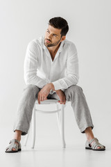 confident man in linen clothes sitting on chair and looking away isolated on grey background