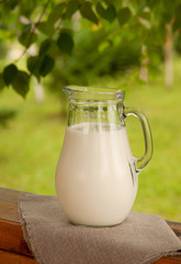 A large jug of milk against the background of green trees. A jug of milk on the windowsill of a wooden pergola. Milk is surrounded by green tree branches.