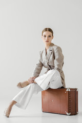 young stylish female tourist sitting on vintage suitcase isolated on grey background