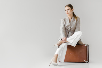stylish woman in linen jacket sitting on vintage suitcase and looking away isolated on grey...