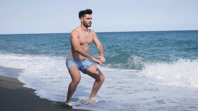 Athletic attractive young man exercising and working out with body weight by the sea, doing squats on sandy beach