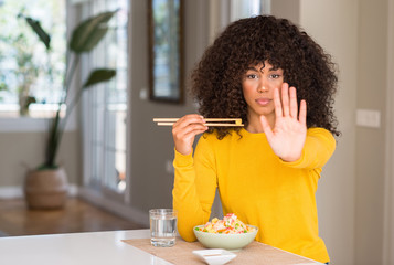 Obraz na płótnie Canvas African american woman eating asian rice at home with open hand doing stop sign with serious and confident expression, defense gesture