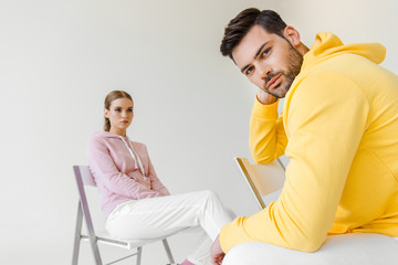 stylish young male and female models in pink and yellow hoodies sitting on chairs isolated on white