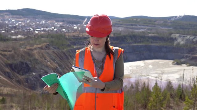 A young woman checks documents against the background of an open-air career on a sunny day. She is wearing an orange vest and a protective helmet.