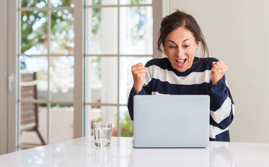 Middle aged woman using laptop at home screaming proud and celebrating victory and success very...
