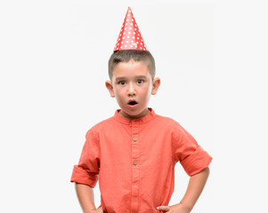 Dark haired little child wearing birthday cap scared in shock with a surprise face, afraid and...