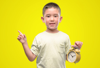 Little child holding alarm clock very happy pointing with hand and finger to the side