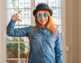 Stylish redhead woman wearing bowler hat and sunglasses smiling and confident gesturing with hand...