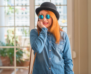 Stylish redhead woman wearing bowler hat and sunglasses looking stressed and nervous with hands on...