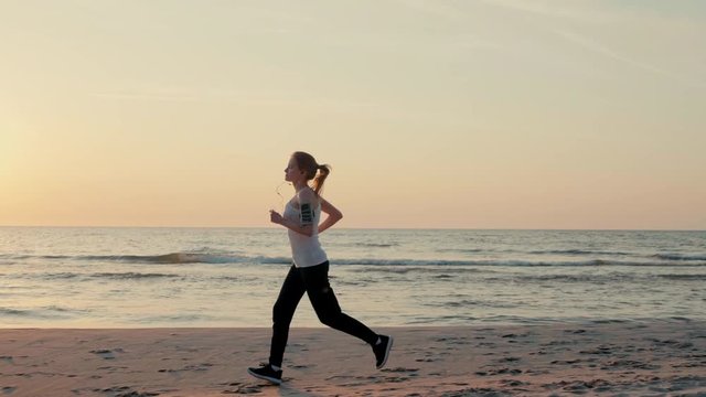 Beautiful Slender Woman is Running along Sea Coast at Sunset and Jogging on Beach with Headphones in SlowMotion