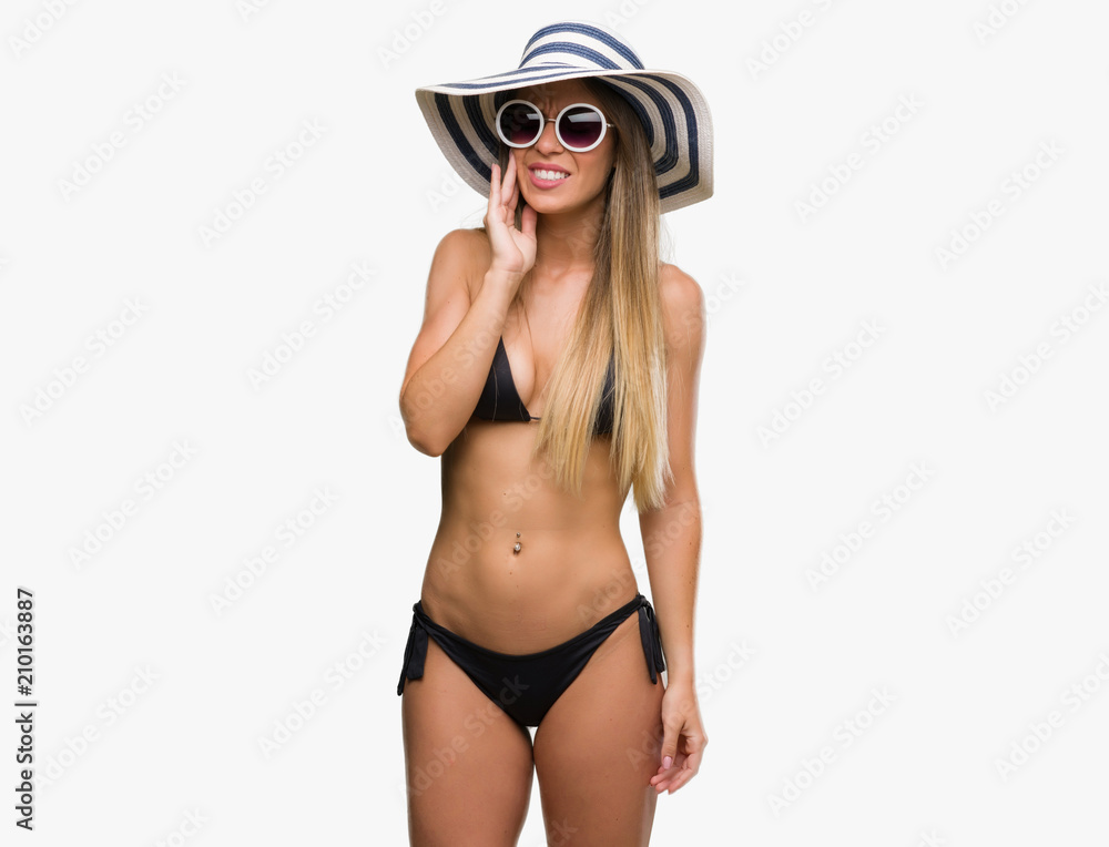 Poster beautiful young woman wearing bikini, sunglasses and hat touching mouth with hand with painful expre