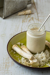 Fresh peeled horseradish roots, a small glass jar with seasoning on a round ceramic green dish on an old wooden table.