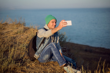 Naklejka na ściany i meble Hipster young man sits and takes a selfie on the brink of break near the sea. Instagram stylization.
