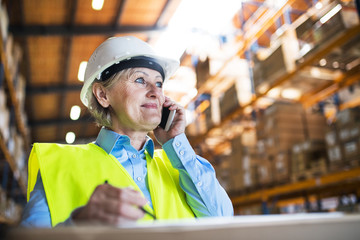 Senior woman warehouse manager or supervisor with smartphone, making phone call.
