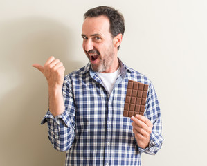 Senior man eating chocolate bar pointing with hand and finger up with happy face smiling
