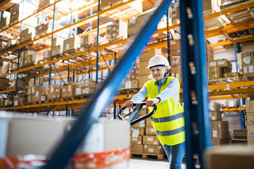 A senior woman warehouse worker pulling a pallet truck with boxes.