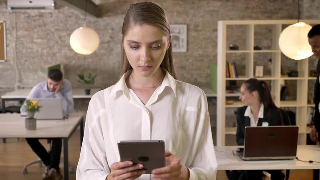 Young concentrated businesswoman is tapping on tablet in office, watching at camera, her colleagues are networking with technologies, working concept, business concept, communication concept