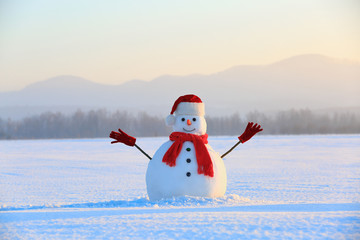 Snowman in red hat and scarf. Christmas scenery. High mountains at the background. Ground covered by snow. Nice cold winter day.