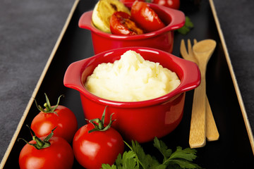 Food is still life. Two red pots with potato puree and fried vegetables on a black tray. Background black marble.