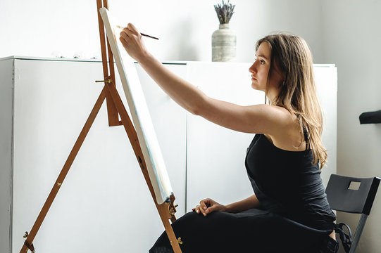 young woman in black clothes and an apron sits in a bright room and paints pictures, brushing oil paints on canvas with malbert