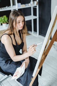 young woman in black clothes and an apron sits in a bright room and paints pictures, brushing oil paints on canvas with malbert