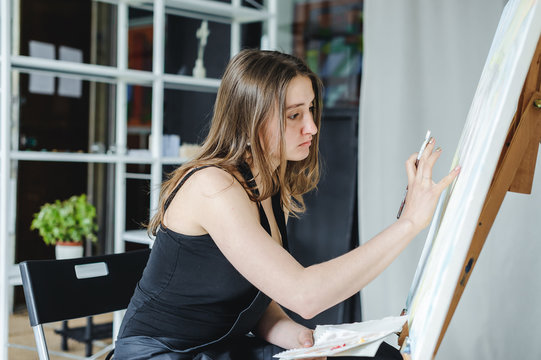 young woman in black clothes and an apron sits in a bright room and paints pictures, brushing oil paints on canvas with malbert