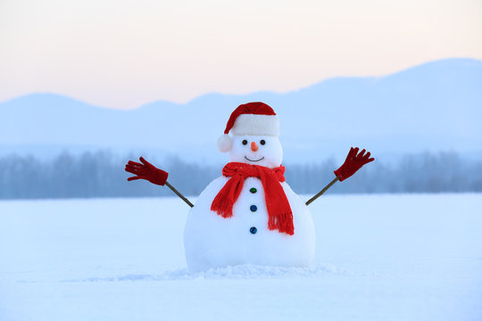 Snowman in red hat and scarf. Christmas scenery. High mountains at the background. Ground covered by snow. Nice cold winter day.