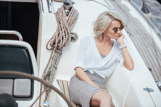 high angle view of pensive blonde girl in sunglasses sitting on yacht and looking away