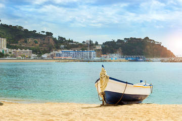Fishing boat moored on the sandy beach on the background of the seaport. Coast of sandy beach and architecture of Spanish beach resort Blanes in summertime. Costa Brava, Catalonia, Spain