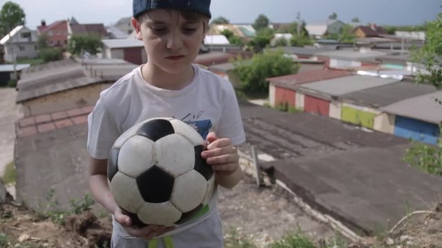 7 year-old Russian white boy in cap with closed eyes and a soccer ball in his hands praying for football team on the background of provinicial Russian urban city garages. Football children education
