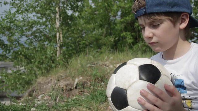 7 year-old Russian white boy in cap with closed eyes and a soccer ball in his hands praying for football team on the background of nature green trees. Football children education concept. Slow motion