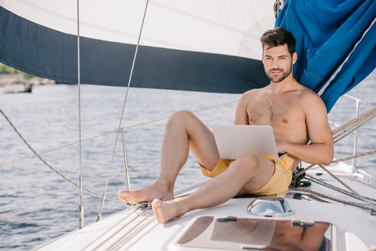 handsome shirtless man in swim trunks using laptop on yacht