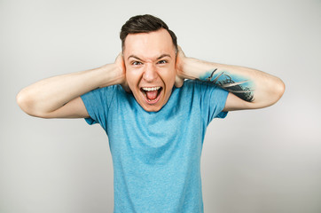 Young guy dressed in a blue t-shirt closing ears on a light background.