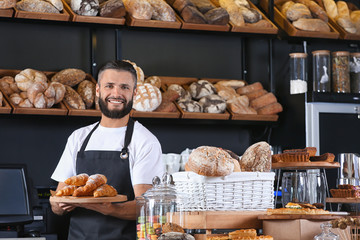 Male baker holding wooden board with delicious croissants in shop - obrazy, fototapety, plakaty