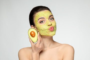 Beautiful young woman with facial mask and fresh avocado on light background
