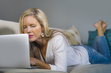 beautiful and happy blond woman early 40s relaxed at home living room using internet on laptop working lying comfortable on sofa at modern apartment