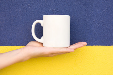 Young Caucasian Woman Holds on Hand Palm Blank Mockup White Mug on Duotone Dark Blue Yellow Painted Wall. Swedish Flag Colors. Template for Text Artwork Lettering. Trendy Minimalist Urban Style.