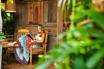 Attractive girl sit on open-air veranda of wooden bungalow with tropical garden view, read romance in paper book. Young woman relaxing in luxury villa on family summer vacation. Countryside lifestyle. - obrazy, fototapety, plakaty