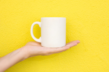 Young Caucasian Woman Holds on Hand Palm Blank Mockup White Mug on Bright Yellow Painted Wall. Airy Breezy Style. Template for Text Artwork Lettering. Trendy Minimalist Urban Atmosphere