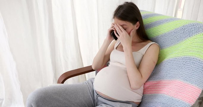 Pregnant woman in expectancy sitting on sofa at home and having phone call looking sad