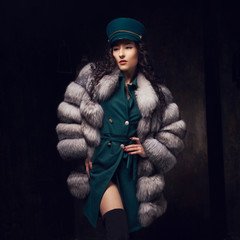 Stylish woman wearing green jacket dress and cap, fur coat and black jackboots posing in dark...