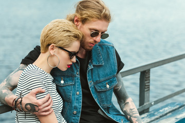 Fototapeta na wymiar boyfriend with tattoos and stylish girlfriend hugging near river and looking down