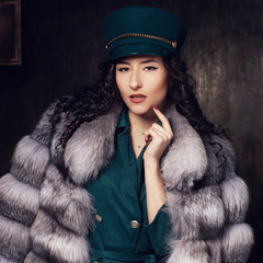 Stylish woman wearing green jacket dress and cap, fur coat and black jackboots posing in dark...