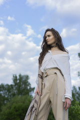 Beautiful stylish girl wearing a trendy white shirt, beige trousers and a pareo poses outdoors against a background of meadow full of wild flowers and blue cloudy sky