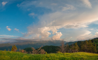 Obraz premium dense evening pink clouds in the sky above the green fields with forest and mountains, picture screensaver