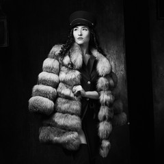 Stylish woman wearing green jacket dress and cap, fur coat and black jackboots posing in dark...