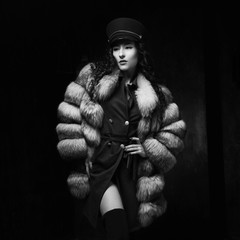 Stylish woman wearing green jacket dress and cap, fur coat and black jackboots posing in dark...
